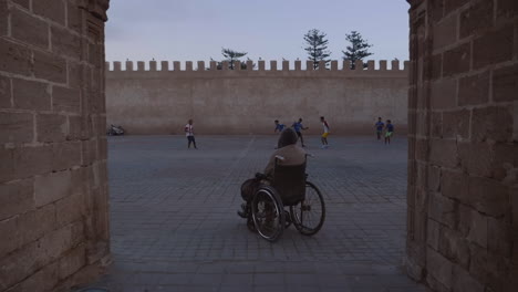 wheelchair-bound man watches young men play soccer football in public square nearby medina in essaouira morocco, handheld shot