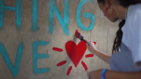 Close-up-cinematic-shot-of-a-white-caucasian-girl-painting-a-red-hearth-and-a-beautiful-quote-on-the-wall-of-an-African-school