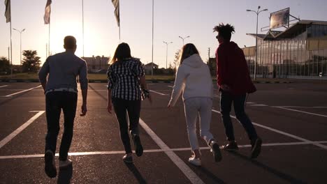 cuatro amigos corren por una zona de estacionamiento vacía al aire libre felicidad divertida, hombres y mujeres jóvenes corren al atardecer y luego saltan juntos libremente