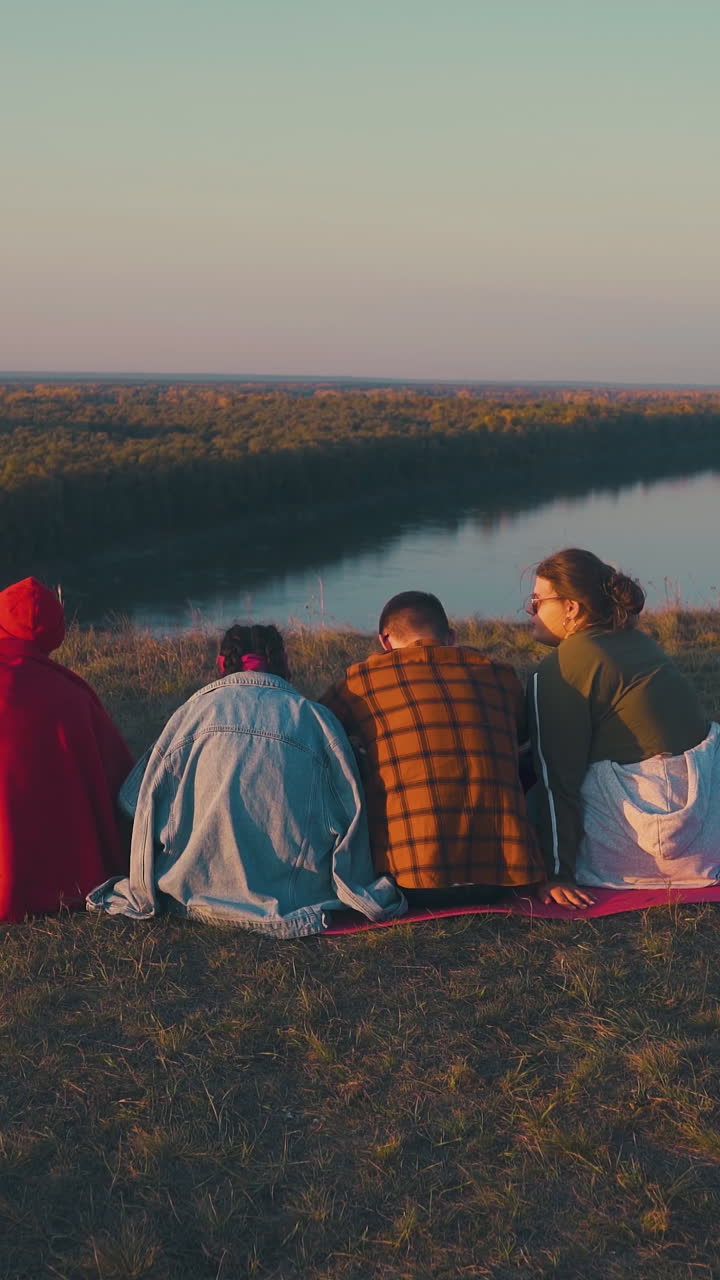 cheerful friends talk at burning bonfire on high river bank in warm autumn evening at sunset backside view