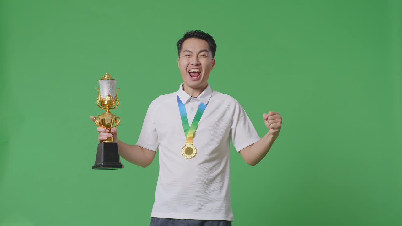 금메달과 트로피를 가진 아시아인 남자가 골을 외치며 스튜디오의 녹색 스크린 배경에서 첫 번째 우승자로 우승을 축하하고 있습니다.