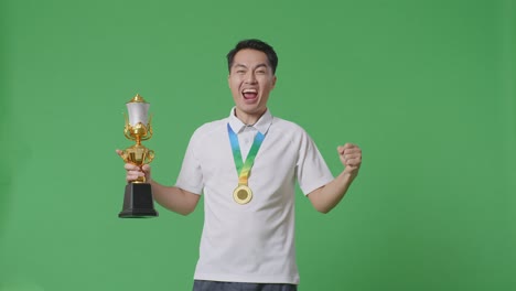 hombre asiático con una medalla de oro y un trofeo gritando gol celebrando la victoria como el primer ganador en el fondo de la pantalla verde en el estudio