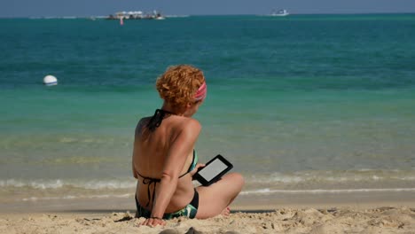 una toma desde atrás de una mujer pelirroja leyendo un libro electrónico en la playa con un bikini
