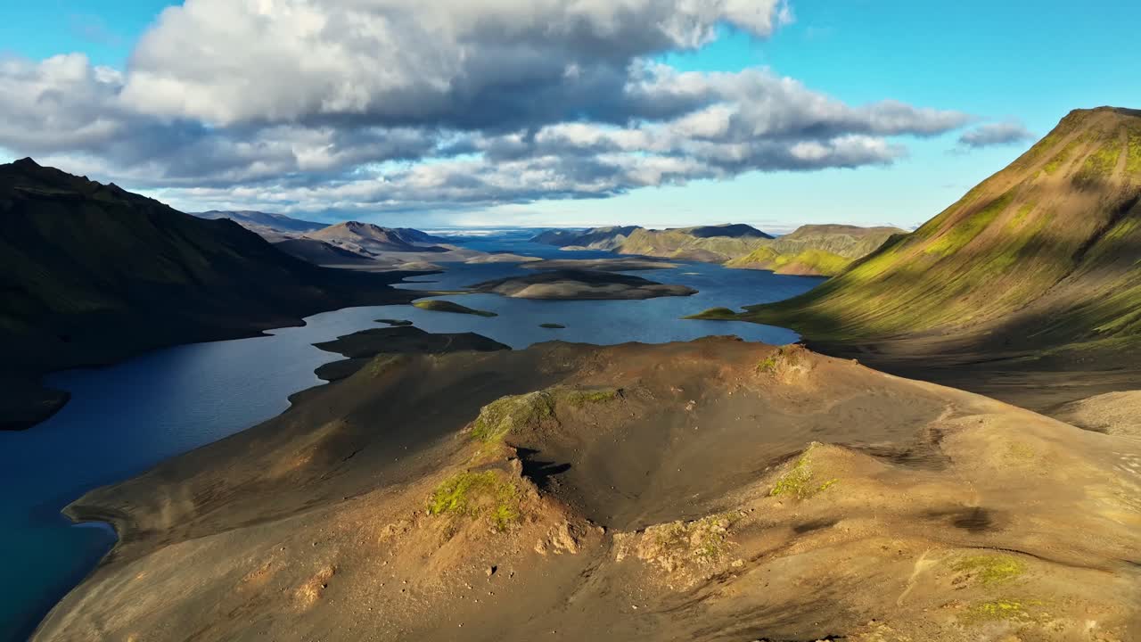 A sweeping aerial view glides above volcanic ridges toward a vast lake system, revealing layered highlands, scattered islands, and dramatic clouds drifting across the Icelandic wilderness