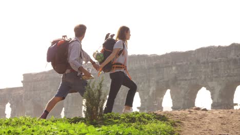 young lovely couple backpackers tourists walking holding hands toward roman aqueduct arches in parco degli acquedotti park ruins in rome on romantic misty sunrise with guitar and sleeping bag slow motion