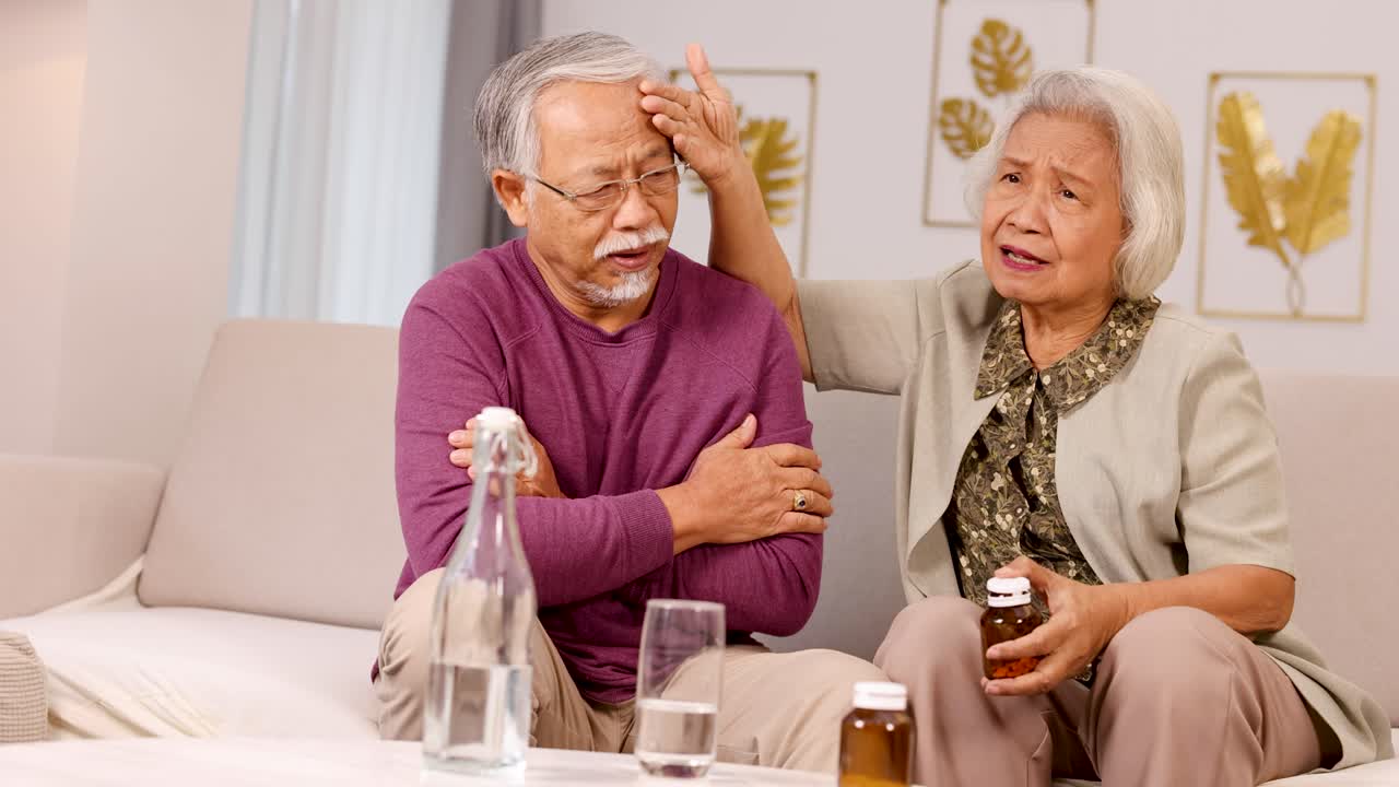 An elderly couple sits on a couch, with one comforting the other who appears unwell. Warm lighting creates a cozy atmosphere