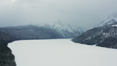 aerial moving over frozen lake with large mountains covered in clouds behind