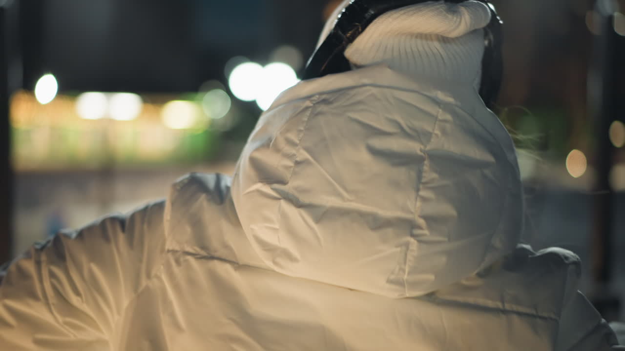 Back view of dancer in white coat and headphones spinning joyfully on snow covered square at night under bright street lamps, arms flowing in movement against urban park backdrop with passersby