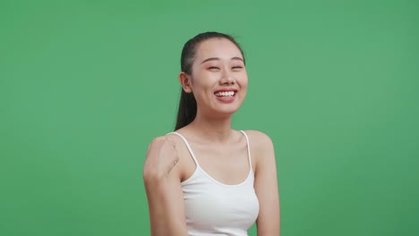 백색 카미솔을 입은 아시아 여성의 측면 초상화. 스튜디오의 녹색 스크린 배경에서 포즈를 취하고 따뜻하게 미소 짓고 있습니다.