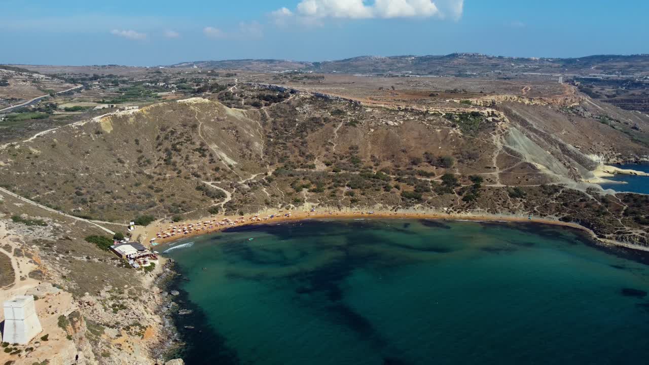 Drone glides over Ghajn Tuffieha Bay, showing cliffs, golden sands, and blue waters, establishing overview at midday