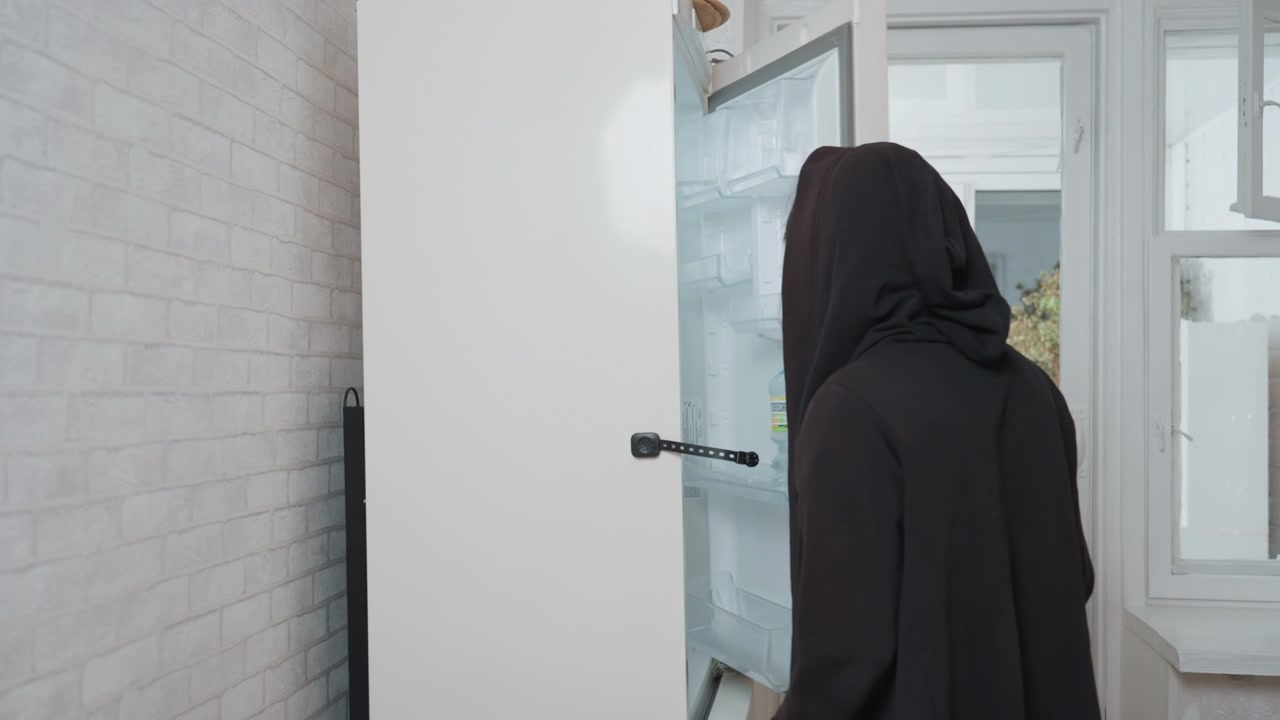 Mutfakta buzdolabını açan, boş rafları inceleyip temizlemeye ve düzenlemeye hazırlanan kişi. Sakin sabah ışığı, modern beyaz fayanslar, huzurlu bir ev işi, minimalist estetik, odaklanmış hareketler.