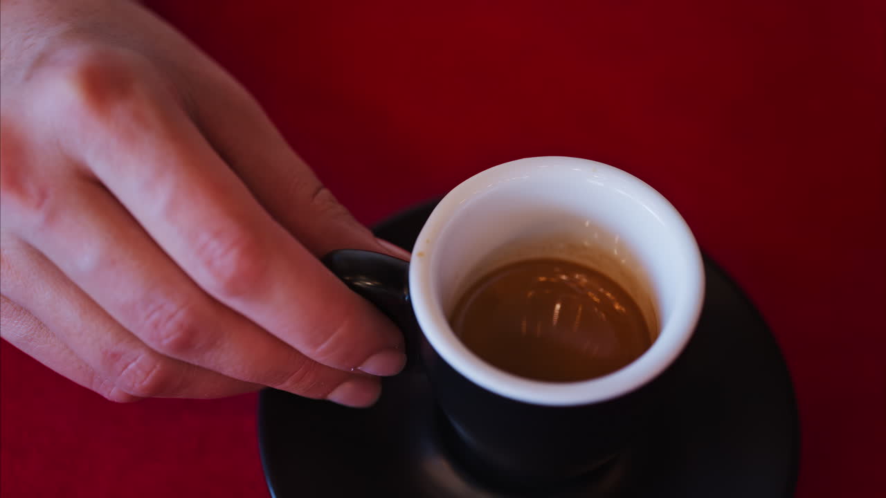 Close up of a woman holding an espresso in a black cup on a red tablecloth at a restaurant