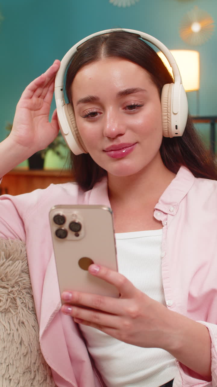 Young woman listening to podcast on sofa wearing headphones nodding while writing insightful notes