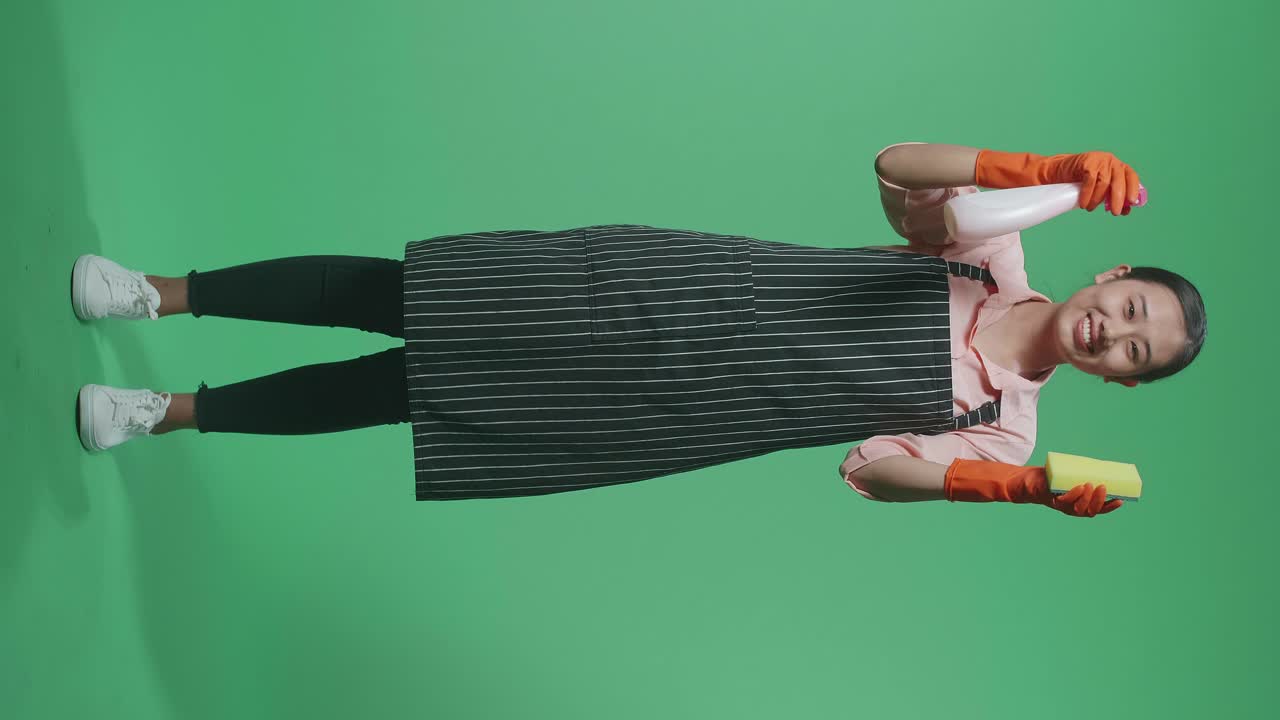 녹색 스크린 스튜디오에 서 있는 동안 스프레이와 부 스폰지를 보여주는 아프론을 입은 미소 짓는 아시아 여성 집사.