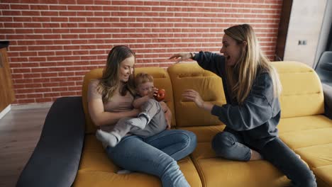 two blonde women play with a child by tickling him on sofa