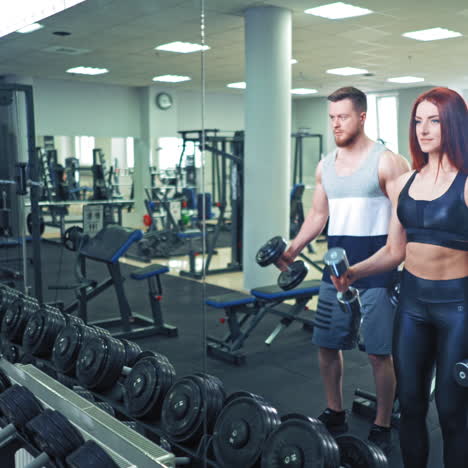 active couple lifting dumbbells together in gym. muscular man and woman holding dumbbells in their hands and doing workout while standing in front of the mirror indoors.