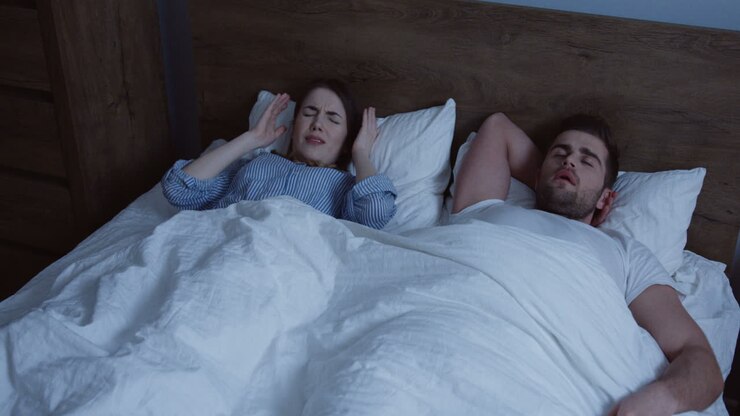 Jonge Kaukasische vrouw kan 's nachts niet slapen omdat haar man naast haar snurkt; de vrouw bedekt haar oren met een kussen.