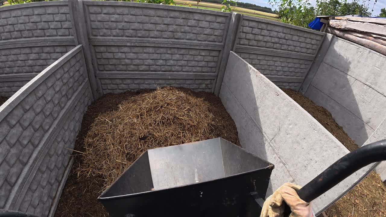 POV shot of farmer emptying wheelbarrow filled with straw and chicken manure onto eco compost heap on sustainable farm