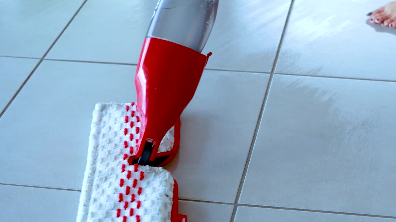 A person uses a red flat spray mop to clean a light-colored tiled floor in a bright indoor setting, with smooth camera movement and natural lighting