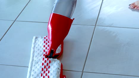 a person uses a red flat spray mop to clean a light-colored tiled floor in a bright indoor setting, with smooth camera movement and natural lighting