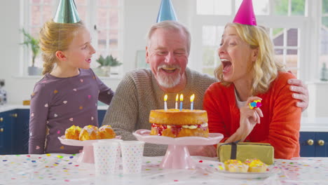 Abuelos-Con-Nieta-Celebrando-Juntos-Soplando-Velas-En-El-Pastel-De-Cumpleaños-En-Casa