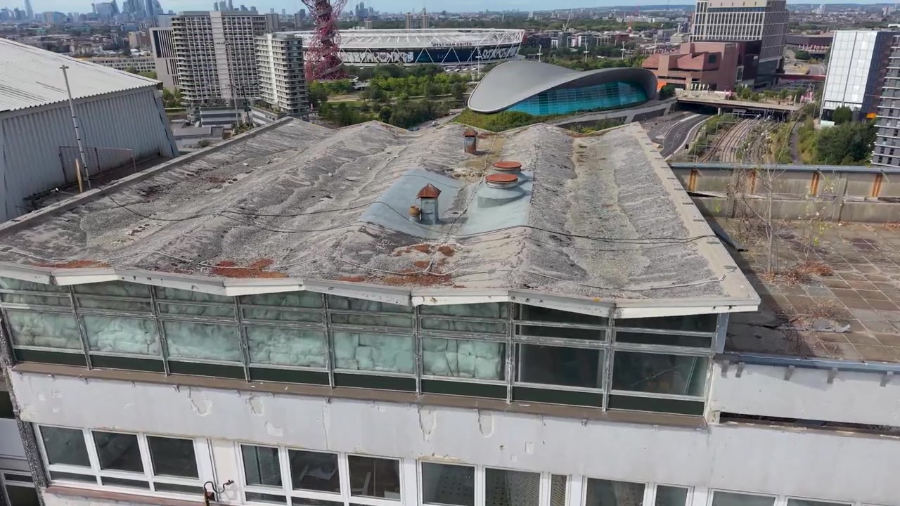 Un dron desciende sobre el tejado de un bloque de torres abandonado en Stratford, pasando por el Centro Acuático de Londres, el ArcelorMittal Orbit y el Estadio del West Ham, luego se aleja y se inclina hacia la fachada.