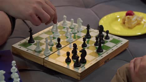 Chess Player Making Move With Black Pawn On Small Chessboard Next To Plate Of Sweet Dessert. closeup shot