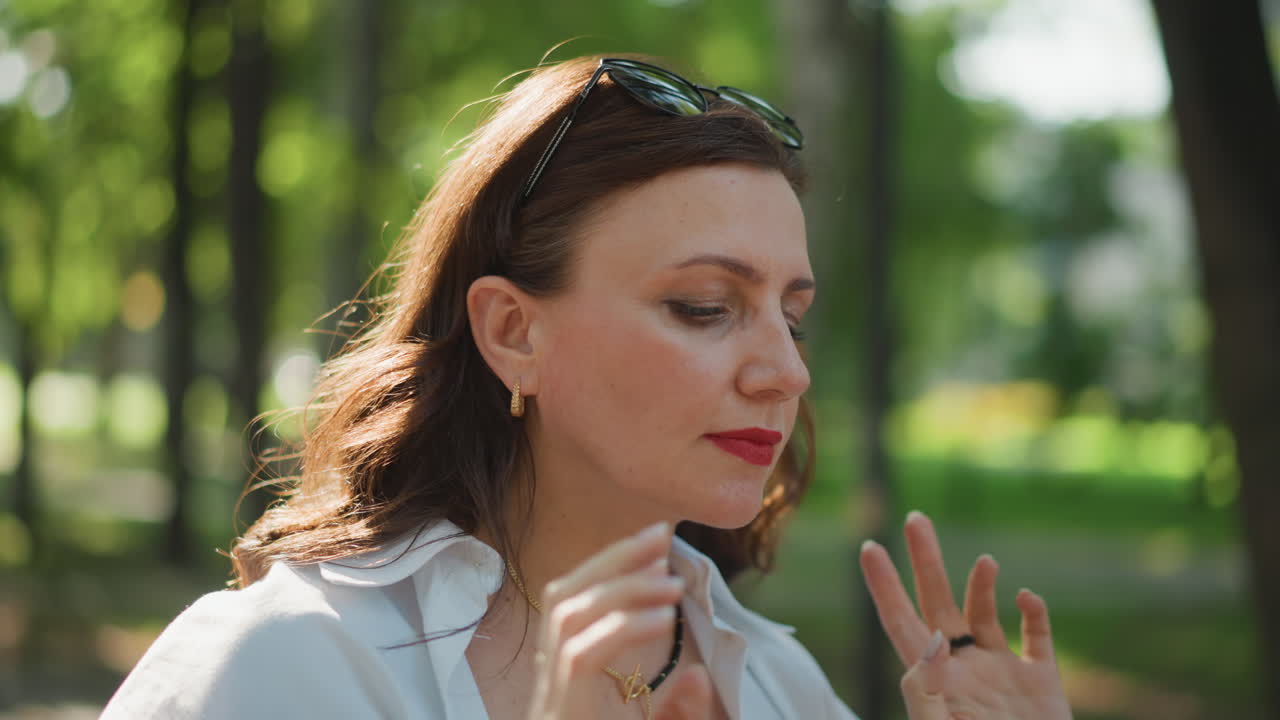 Donna caucasica che si sistema gli occhiali da sole al parco, ritratto di moda ravvicinato, rossetto rosso e sguardo sicuro, bokeh retroilluminato e ombra urbana, umore assertivo, focus sull'accessorio chic, tono da dichiarazione di stile di vita