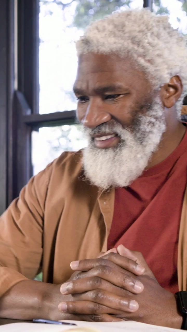 Vertical video: Noticing line, senior man leaning over notes by window, picking pen, rust shirt