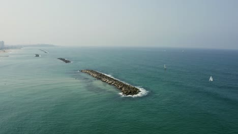 Blue-Waves-By-The-Mediterranean-Sea-Splashing-On-The-Rocks-On-The-Seawalls-With-Sailing-Boats-Near-The-Tel-Aviv-Beach-In-Tel-Aviv,-Israel