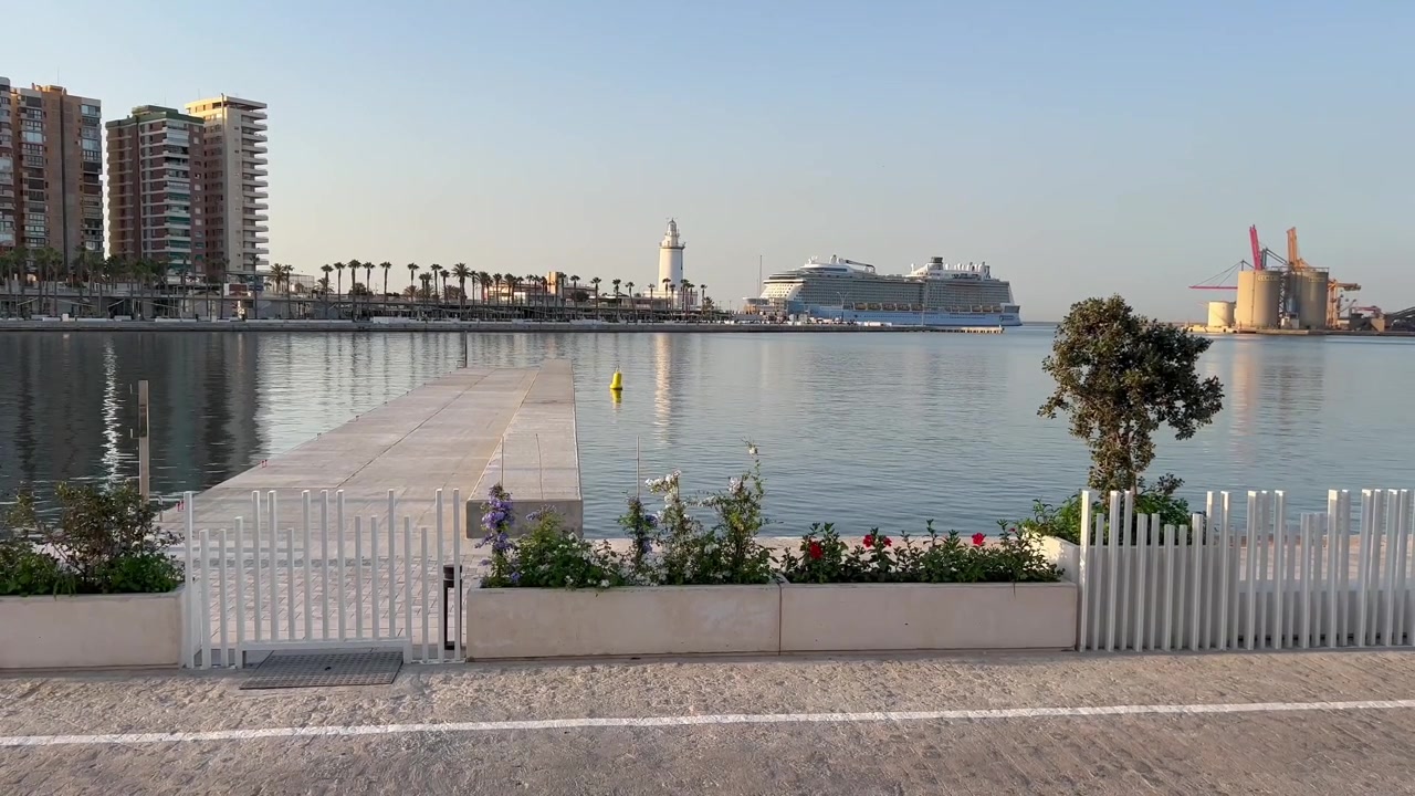 Static ground-level shot of Malaga harbor at golden hour. Cruise ship, lighthouse, and city skyline reflect on calm water.