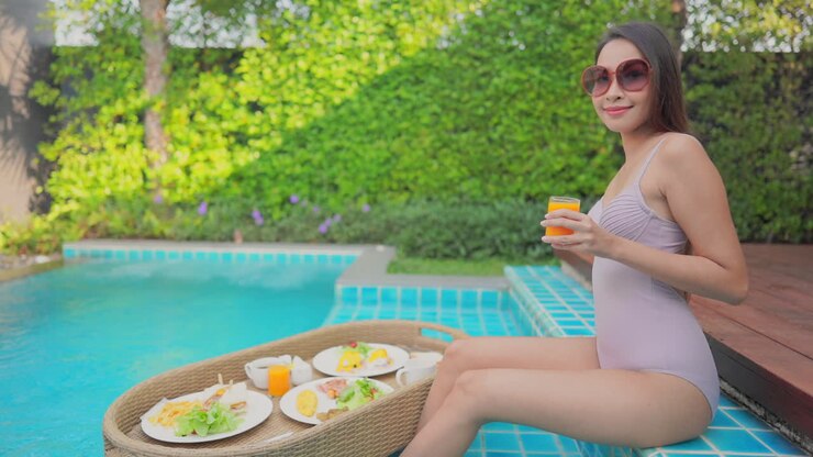 Young woman sitting on the poolside step holds an orange juice in one hand as a service platter full of breakfast foods floats in the pool by her feet