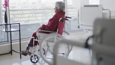anciana paciente biracial en silla de ruedas mirando por la ventana en la sala del hospital, cámara lenta