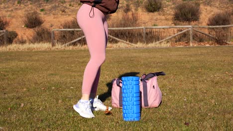 una mujer joven con ropa deportiva coloca su bolsa en la hierba y comienza a masajear sus piernas con un rodillo de espuma