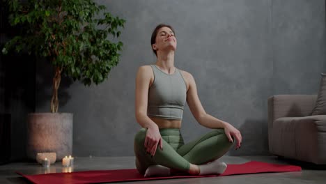 Confident-and-calm-brunette-girl-in-a-gray-top-and-green-sweatpants-sits-in-the-lotus-position-on-the-red-carpet-and-does-sports-does-a-neck-warm-up-near-lit-candles-and-a-houseplant-in-a-modern-apartment