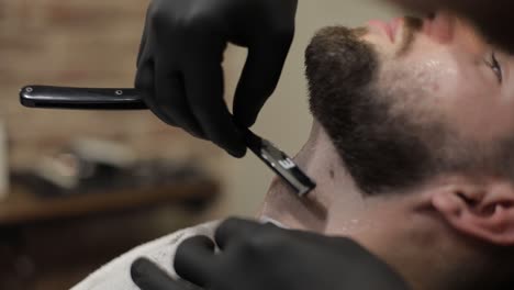 Client-with-black-beard-during-beard-shaving-in-barber-shop.-Groom,-masculine