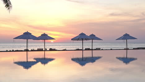 Colorful-sunset-along-with-beach-umbrellas-reflect-in-a-resort-swimming-pool