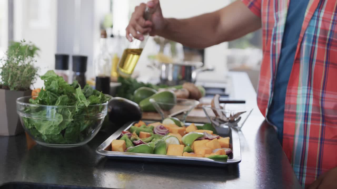 sección media de un hombre biracial preparando comida, vertiendo aceite en las verduras en una cocina soleada, cámara lenta