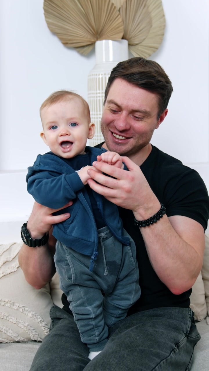 Man in black t-shirt sits on the sofa holding his son. Little kid smiles to camera, dad kisses and hugs his adorable baby. Vertical video