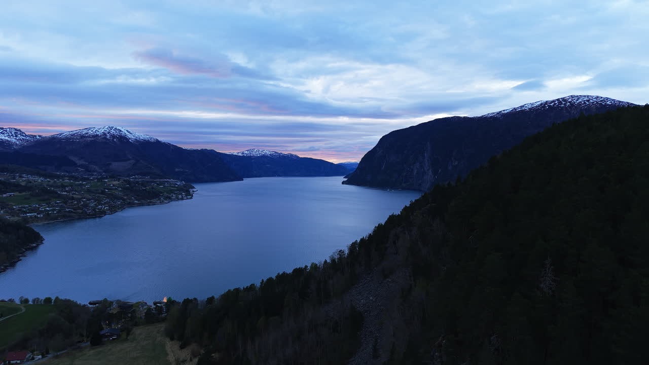 Senja menyelimuti fjord Norwegia dengan cahaya lembut, dikelilingi oleh pegunungan bersalju dan hutan lebat. Ideal untuk fitur perjalanan atau dokumenter alam.