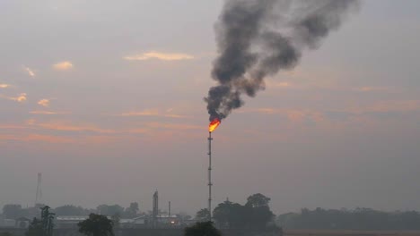 Petrochemical-refinery-flaming-smoking-flare-stack-rising-over-misty-agricultural-farmland-at-sunrise