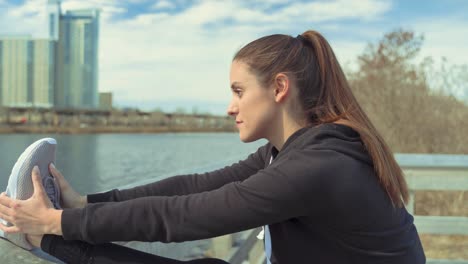 Fitte-Und-Flexible-Junge-Frau,-Die-Sich-Vor-Dem-Morgendlichen-Joggen-Das-Bein-Gegen-Einen-Holzzaun-Am-Wasser-Streckt,-Während-Sie-Konzentriert-Und-Entschlossen-Aussieht