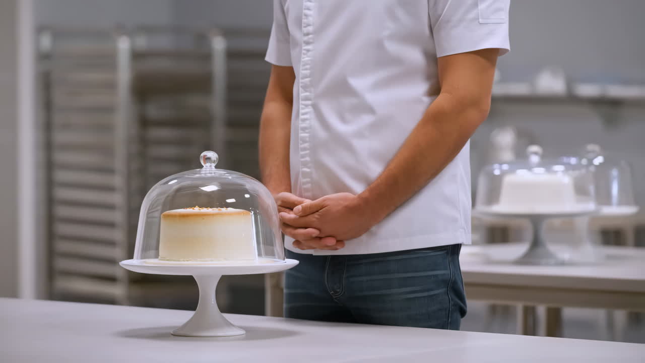 A chef stands next to a cake under a cloche in a bakery setting