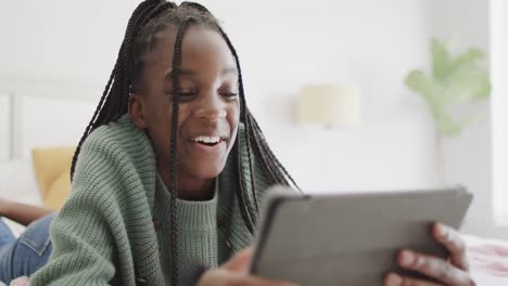 Happy-african-american-teenage-girl-lying-on-bed-using-tablet-and-laughing