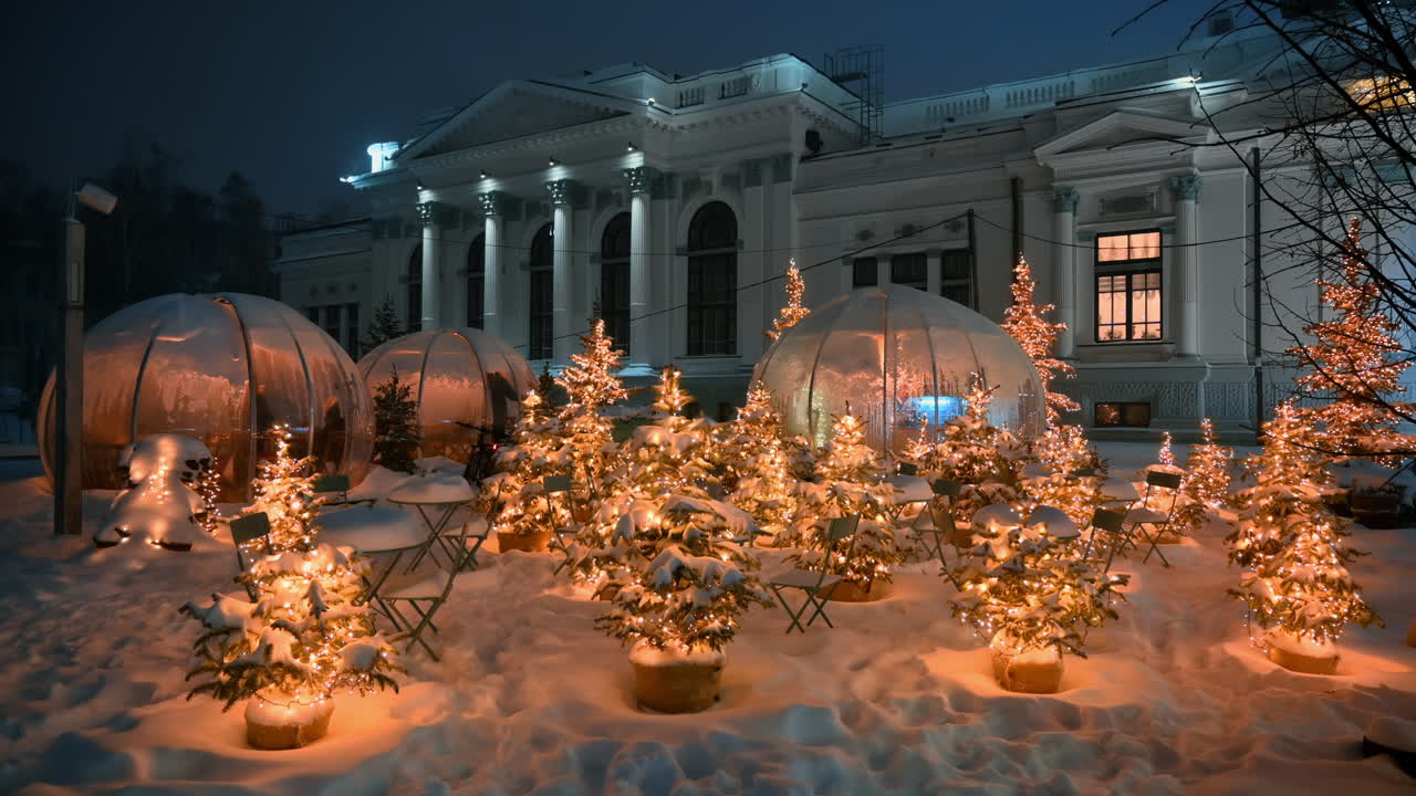 Christmas trees with garland lamp lights near coffee tables during snowfall. Outdoor coffee shop. Winter evening in Chisinau, Moldova