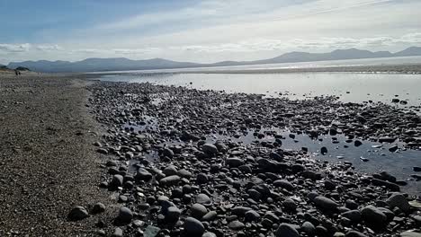 Hazy-Snowdonia-mountain-range-across-idyllic-slow-motion-across-shimmering-Irish-Seascape-from-Newborough-rocky-beach-shoreline