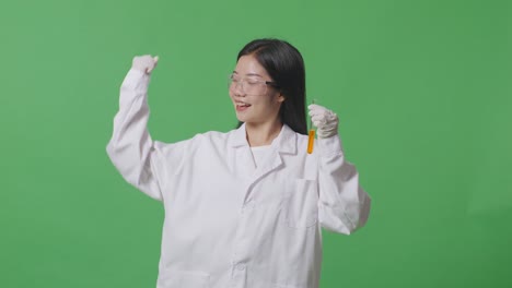 asian woman scientist with orange liquid in the test tube smiling and flexing her bicep while standing on the green screen background in the laboratory