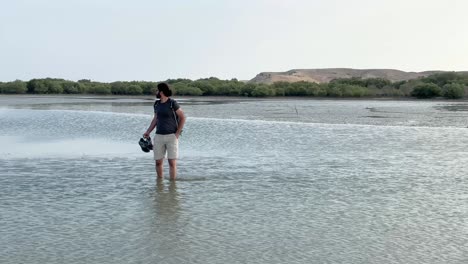 A-middle-eastern-man-is-walking-bare-foot-grab-shoes-boot-in-the-sea-beach-day-time-in-Qatar-Doha-Arabian-Gulf-tourism-hiking-tidal-road-mangrove-mangroove-forest-stream-green-leaves-tree-background