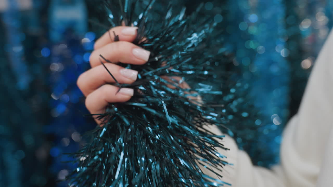 Close-up of person feeling texture of dark blue shimmering tinsel, enjoying tactile experience in beautifully decorated store, with blurred festive decor in background