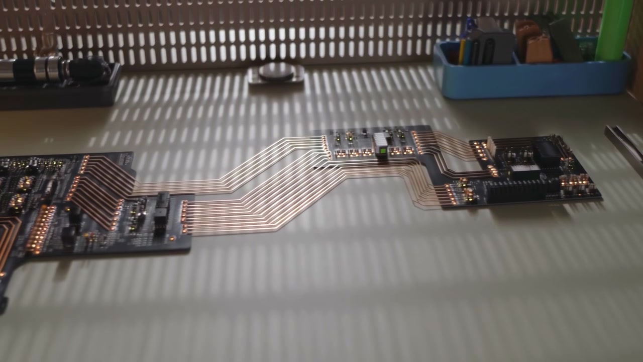 La telecamera si muove attraverso schede elettroniche interconnesse e percorsi conduttivi esposti su un tavolo, mentre una forte luce diurna a strisce proveniente dalle tende della finestra crea ombre ripetute.